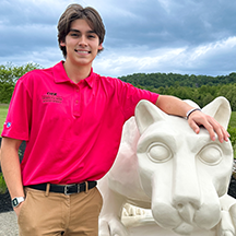 PSU-LV male student in bright pink New Student Orientation leader shirt smiling, standing next to the PSU-LV lion shrine.