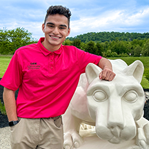 PSU-LV male student in bright pink New Student Orientation leader shirt smiling, standing next to the PSU-LV lion shrine.